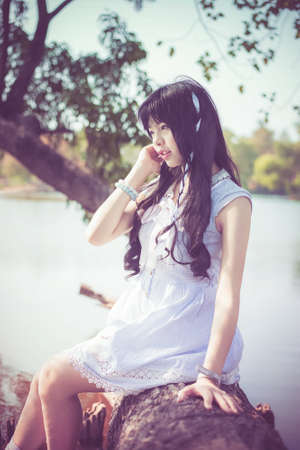 A cute Asian Thai girl is sitting on a tree trunk on the riverside with soft breeze blowingの写真素材