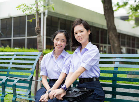 Cute Asian Thai high schoolgirls student couple in school uniform sit wih a happy smile face together on a benchの写真素材