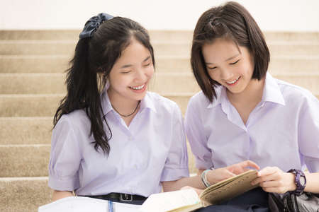 Cute Asian Thai high schoolgirls student couple in school uniform sit on the stairway discussing homework or exam with a happy smiling face together on a building stairsの写真素材