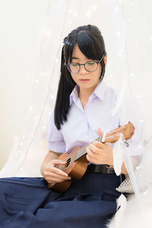 Cute Asian Thai high school girl in uniform and glasses sitting and playing guitar in a soft cloth curtain decorate with feather and light in schoolgirl fashion and music recreation education concept in white studio background.の写真素材