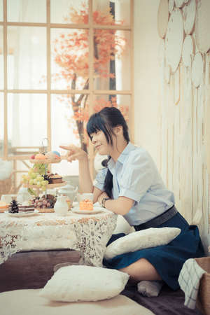 Cute Asian Thai high school girl in uniform is playing with dessert food decoration with various food fruit and candy in a lovely cafe or retaurant scene in vintage color style.の写真素材