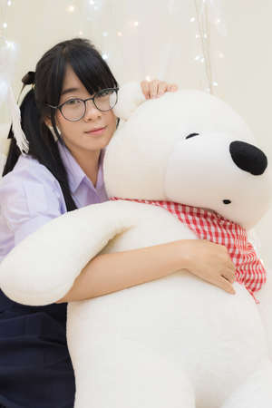Cute Asian Thai high school girl in uniform and glasses sitting and hugging fluffy teddy bear doll a soft cloth curtain decorate with feather and light in schoolgirl fashion recreation concept.の写真素材