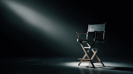 A director's chair stands alone against a dark backdrop, highlighted by a strong overhead spotlight. The wooden frame and canvas seat create a stark contrast with the deep shadows. The composition uses dramatic lighting and negative space suggesting anticipation, waiting, and readiness for commercial or editorial use.の素材