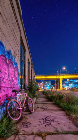 A bicycle rests against a brick wall adorned with colorful graffiti. The composition includes a walkway, street lights, and an elevated road structure, under a deep blue night sky. The image conveys an urban atmosphere with potential uses for design, editorial content, and commercial applications.の素材