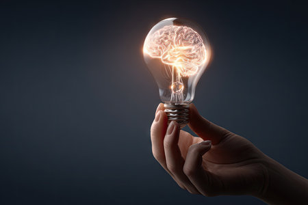 A human hand gently holds a light bulb, revealing a glowing brain inside. The image features a dark background with a focus on the illuminated object. The lighting is soft, creating a sense of depth and intellectual curiosity. This composition is suitable for use in conceptual projects related to ideas, innovation, or education.の素材