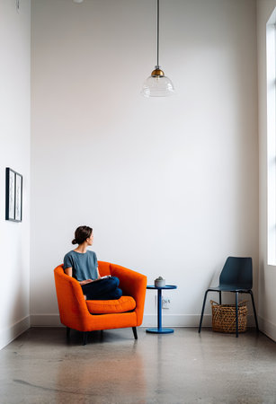 A woman is seated in an orange armchair, looking out of a window. The scene features a minimalist interior with white walls, a gray floor, and a single hanging light. The composition is clean, with the main subject in focus, and could be utilized for various commercial and editorial projects.の素材