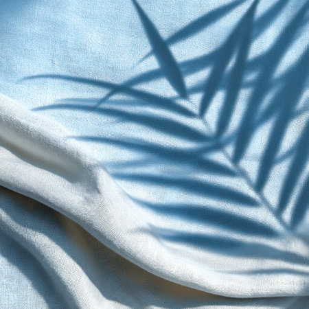 An overhead view reveals a shadow of a palm leaf on a textured white fabric. The play of light and shadow creates a contrast of blue against white. This image evokes feelings of summer and relaxation. Suitable for use in lifestyle or decorative projects.の素材