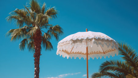 An image captures a white parasol with an ornate design and a palm tree set against a vivid blue sky. The composition emphasizes the contrast between the textures of the natural elements and the manufactured object. This image may be suitable for various uses, including travel, lifestyle, or decorative projects.の素材