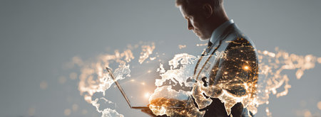 A man in a suit looks at a laptop, a map of the world is projected onto the image creating a composite effect. The image incorporates light and dark shades. It has a high-key composition with a focus on business and technology, potentially suitable for editorial and commercial applications.の素材