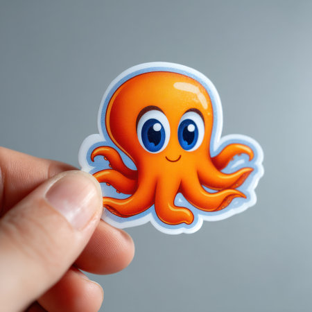 A close-up view displays a hand holding an octopus sticker against a plain background. The cartoon octopus features bright orange and red hues with blue eyes. The illustration uses a clean, graphic style, likely suitable for children's designs or educational material and potential commercial applications.の素材