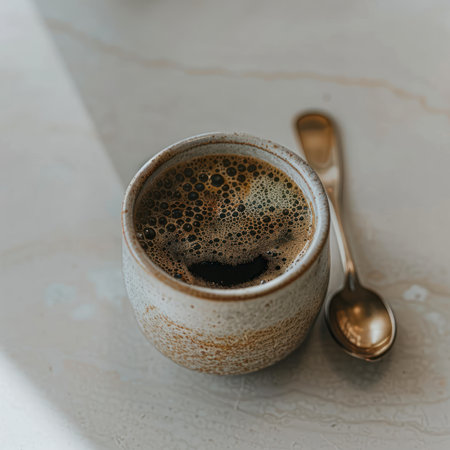 A close-up shot presents a cup filled with coffee, alongside a spoon, resting on a textured surface. The image highlights the cup's texture and the dark coffee color, with overhead lighting. Suitable for commercial or editorial applications, the photo could be used for various projects.の素材