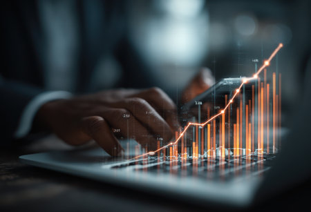 A close-up shot showcases hands interacting with a laptop and a smartphone. An illuminated financial graph, rendered in vibrant orange, overlays the scene. The image displays a dark aesthetic with shallow depth of field, possibly taken indoors. This visual could be utilized for economic, business, or technology related content.の素材
