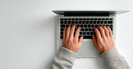 An overhead view displays hands interacting with a laptop keyboard. The image shows a minimalistic composition, featuring a laptop on a white surface. The neutral color palette and simple aesthetic suggest a clean workspace environment suitable for various commercial or editorial applications related to technology or work.の素材