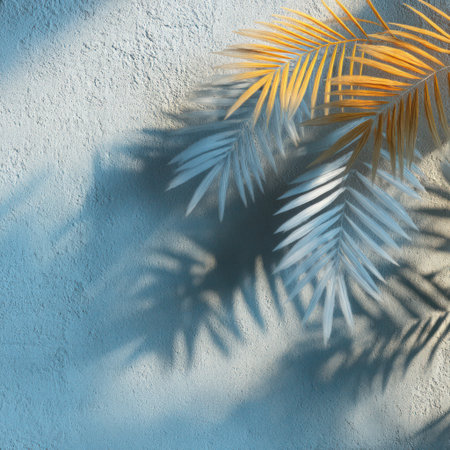 Palm fronds with golden and silver hues are presented against a rough, light blue wall. The composition highlights the shadows cast by the leaves, creating a pattern. The scene is bathed in warm sunlight, enhancing the contrast between light and shadow. Ideal for various commercial uses.の素材