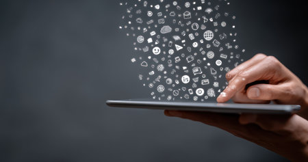 A person's hands interact with a tablet device, as numerous digital icons appear to emanate from the screen. The image showcases a dark background with an overhead perspective. The composition features a neutral palette, suggesting a technological or informational context, appropriate for business or editorial applications.の素材
