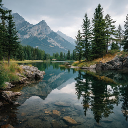 A serene landscape showcases a tranquil lake mirroring towering evergreen trees and rugged mountain peaks. The composition features soft lighting with a palette of greens, blues, and grays. This natural scene could be used for travel, environmental, or promotional visuals.の素材