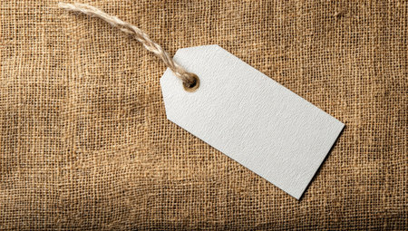 A clean white tag hangs suspended by a natural string against a textured brown textile background. The composition features overhead lighting and highlights the blank space available on the tag. It's suitable for various commercial uses, including product labeling and design projects. The burlap adds a rustic and tactile element.の素材