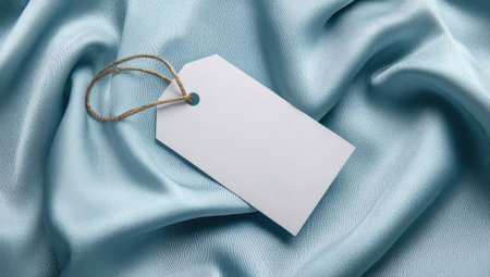 A white price tag hangs over a blue silk fabric. The fabric exhibits elegant folds and a soft sheen. The lighting is diffused, enhancing the texture of the material. This image is suitable for promotional materials, advertising, or presentations, offering ample space for text.の素材