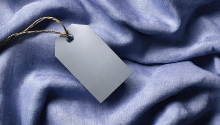 A close-up view displays a white label with a string attached, positioned diagonally against a textured, light blue fabric. The soft focus and gentle folds of the textile create a visually appealing backdrop. Suitable for presentations or advertising projects requiring a clean and neutral element, with potential for showcasing product pricing or branding information.の素材