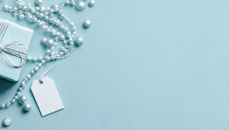 A top-down view showcases a pale blue gift box adorned with a silver ribbon, alongside a lustrous pearl necklace and a blank white tag. The elements rest on a light blue backdrop, offering a clean, airy aesthetic. This image is suitable for commercial purposes, including advertising and editorial use in fashion or lifestyle contexts.の素材