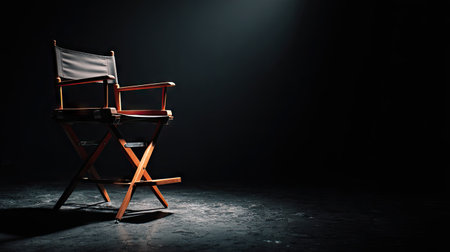 A wooden director's chair stands centered on a dark stage, illuminated by a single overhead light. The chair's brown wooden frame and black seat contrast against the shadowed background. This artistic composition suggests themes of performance, cinema, or creative direction, potentially suitable for various commercial or editorial uses.の素材