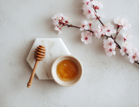 A wooden honey dipper rests beside a small bowl of honey and a branch of delicate pink blossoms. The composition is set against a plain white background, with soft natural light highlighting the textures. This image has potential uses in articles or marketing material related to wellness or food.の素材