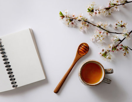 A close-up captures a wooden honey dipper resting beside a cup of tea, a notebook, and delicate white blossoms. The composition features a neutral white background. The soft lighting and high-angle perspective contribute to a calm, inviting aesthetic. Suitable for culinary, lifestyle, or wellness-related content, it is ideal for various commercial uses.の素材