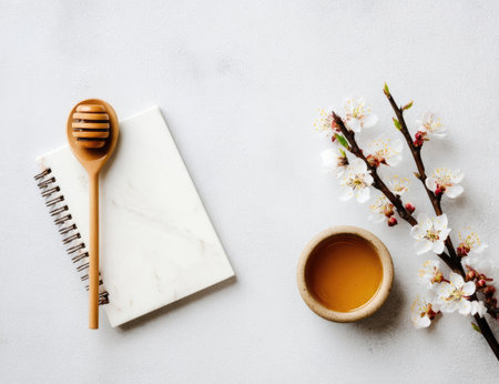 An overhead shot showcases a wooden honey dipper, a blank notepad, a small bowl of honey, and flowering tree branches. The composition exhibits a minimalist aesthetic with soft lighting and a clean white background. This image could be used for food, wellness, or lifestyle purposes.の素材