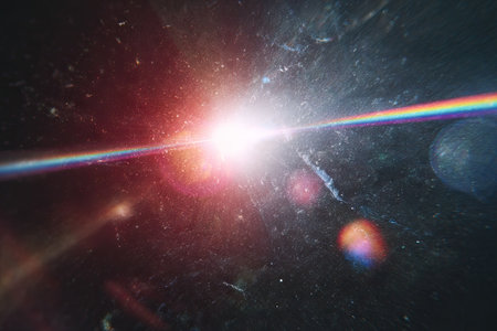 This image features a bright light flare with a rainbow effect, set against a dark, blurred background with particle-like elements. The composition showcases a dynamic burst of light, with a variety of colors and textures. Suitable for illustrating concepts related to energy, abstract art, or digital design, this image is ideal for commercial use.の素材