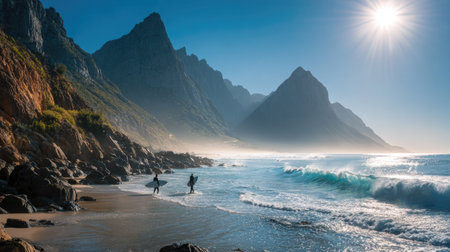 Two figures with surfboards stand near the ocean's edge, framed by towering mountains and the dynamic flow of waves. The scene presents a vivid color palette, with a bright sun overhead and clear, blue skies meeting the sea. This landscape suggests outdoor recreation, appropriate for travel or adventure content.の素材