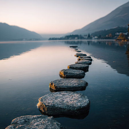 An image captures a series of stepping stones stretching across tranquil water. The stones lead towards a mountain range, all bathed in the soft light of either dawn or dusk. The composition highlights reflections with a smooth surface. Suitable for editorial projects and commercial applications, evoking a sense of peace.の素材
