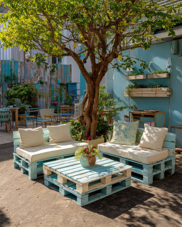 This image presents an outdoor seating area composed of pallet furniture. The scene features light-colored cushions and a wooden coffee table, all set against a background with blue walls and a lush tree. The daylight suggests a relaxed and inviting atmosphere, potentially suitable for commercial or editorial applications.の素材
