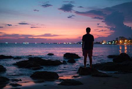 A man stands silhouetted on a beach, gazing at a vibrant sunset over the ocean. The image showcases soft colors of the sky, reflected in the water. The composition highlights the figure in a moment of contemplation. This image could be suitable for various uses, including editorial and commercial projects.の素材