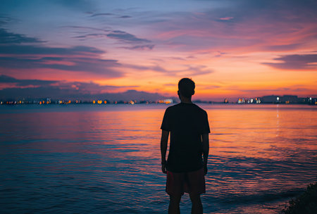A person stands silhouetted against a colorful sunset over a body of water. The sky displays vibrant hues of orange, pink, and blue. The scene is bathed in soft, natural light, creating a tranquil atmosphere. This image could be suitable for various commercial and editorial applications, emphasizing serenity and contemplation.の素材