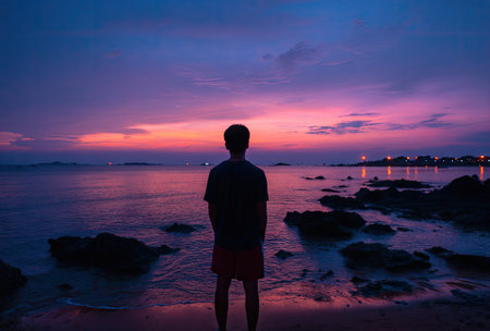 A person stands silhouetted against a colorful sunset over a tranquil ocean. The image showcases the back of the individual in front of the water and sky. The scene is bathed in shades of purple and orange, suggesting a serene outdoor setting. Suitable for various editorial and commercial applications.の素材
