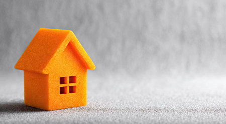 An orange miniature house is the main subject against a gray textured backdrop. The image features a shallow depth of field, with the house in sharp focus. The lighting appears soft and diffused, creating subtle shadows. This image could be used for various design projects, illustrating concepts of home ownership or real estate.の素材