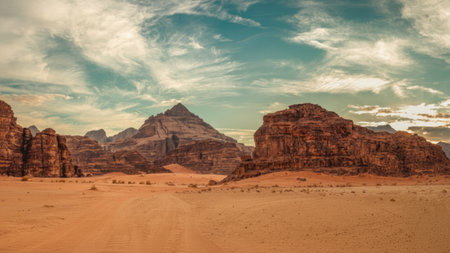 The image features a vast desert scene dominated by imposing rock formations. The composition showcases a warm color palette with shades of brown and orange in the foreground and the majestic rocks. The sky presents a mix of blue and white, suggesting a daytime setting. Suitable for various commercial and editorial applications.の素材