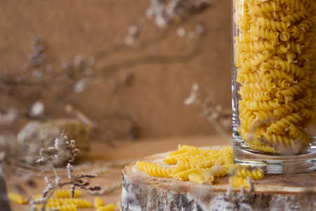 Spiral pasta in a glass jar on a wooden stand, next to it are dry tree branches, on a beige...