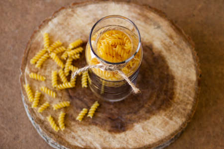 Spiral pasta in a glass jar on a wooden stand, scattered on the table. High quality photoの写真素材