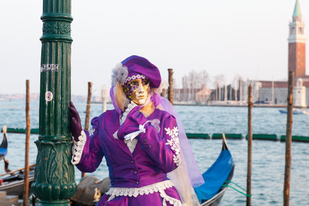 Venice 2017 Carnival, Italy. Woman in classical purple mask and costume posing in front of gondolas at the lagoon at Pizza san marco with San Giorgio Maggiorre in the backgroundのeditorial素材