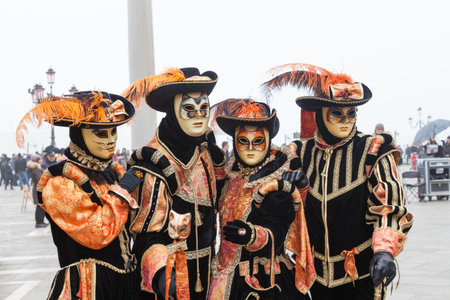 Feb 2017 Venice Carnival, Italy. group of four people in orange and black cat costumes and masks in classic style on a rainy misty Piazzo San Marco with copy space and blurred crowd in the distanceのeditorial素材