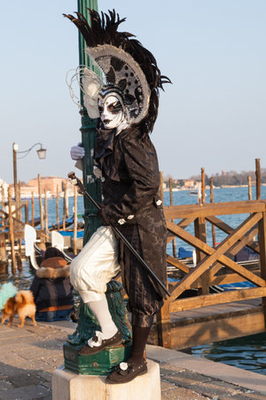 2017 Venice Carnival, Veneto, Italy. Man in a black and white costume and mask with headdress at the lagoon at Piazza San Marco with gondolas  and wooden jetties behind standing full length on a pole .のeditorial素材