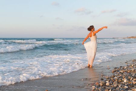 Barefoot young woman in a long white partially wet dress, enjoys a lonesome walk in the water, on a sandy beach in a late summer day, at dusk.の写真素材