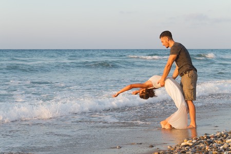 Loving young couple at the beach , in a late summer hazy day at dusk, wearing  a white dress and shorts, enjoying, going barefoot in the ocean water, getting wet, teasing and kissing one another.の写真素材