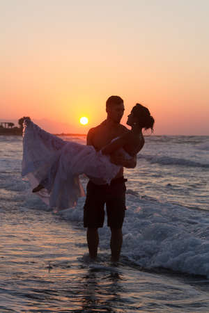 Just married young couple at the beach , in a late summer hazy day at dusk, wearing  a wedding dress and shorts, enjoying walking barefoot, getting wet, teasing and kissing one another.の写真素材