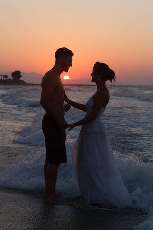 Just married young couple at the beach , in a late summer hazy day at dusk, wearing  a wedding dress and shorts, enjoying walking barefoot, getting wet, teasing and kissing one another.の写真素材