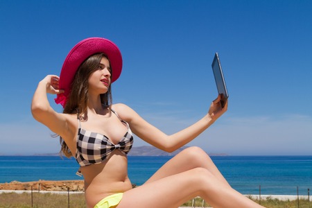 Gorgeous young busty naughty long haired brunette teen girl in swimwear enjoying the sun on a fresh spring morning by the ocean, poses for selfies using her tablet.の写真素材