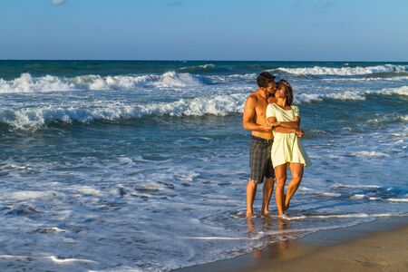 Happy attractive young couple in beachwear, enjoying a summer dusk at the beach, having fun walking barefoot, getting wet, kissing and teasing one another.の写真素材