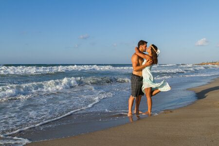 Loving young couple at the beach in a late summer hazy day at dusk, wearing a turquoise dress and shorts, enjoying  going barefoot in the ocean water, getting wet, teasing and kissing one another.の写真素材