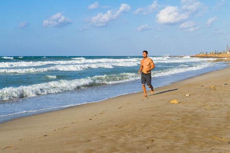 Young man in his twenties jogging along the shoreline on a sandy beach, late afternoon in the summer.の写真素材